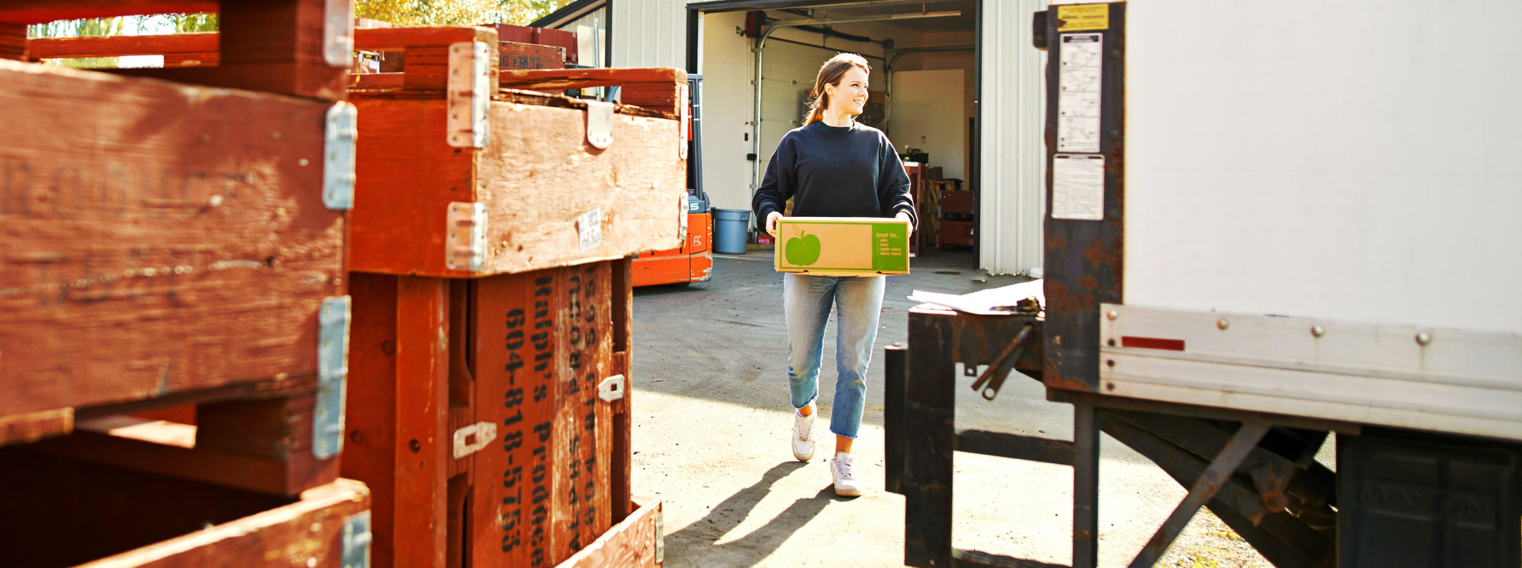 A smiling woman carries a box of produce from a warehouse toward a truck.