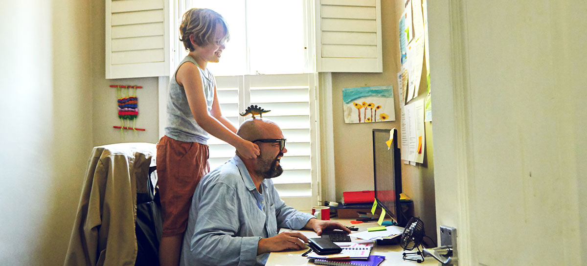 Father sitting at computer desk with a dinosaur toy on his head, with son sitting on his chair behind him pulling his ears