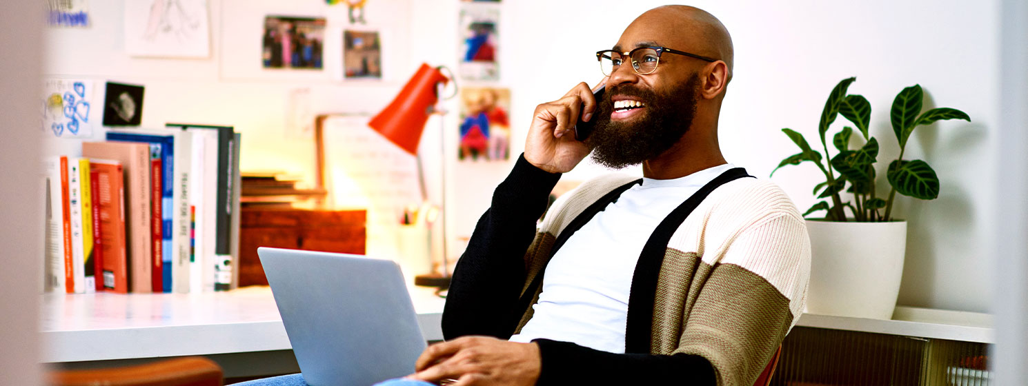 Dark skinned man, in his office, leans back in a chair with a laptop. He’s on the phone and smiling.