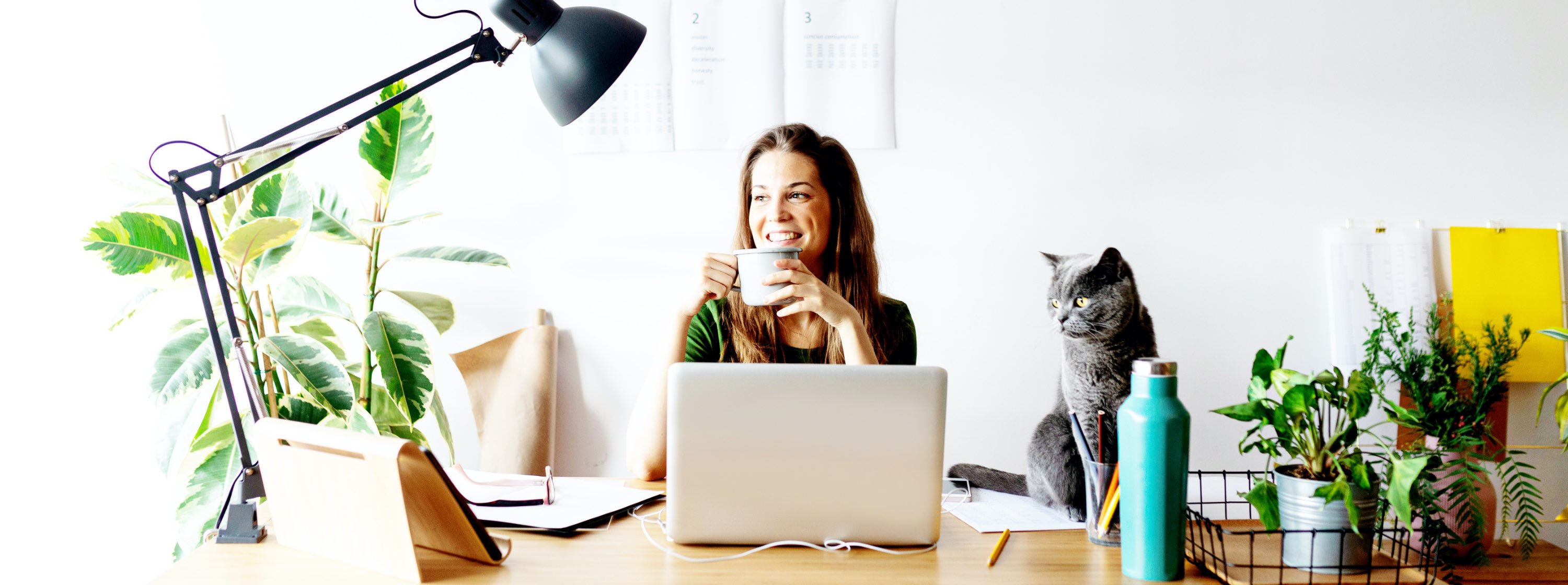 smiling woman sitting at her home office desk, holding a coffee mug, surrounded by plants with her cat on the desk beside her
