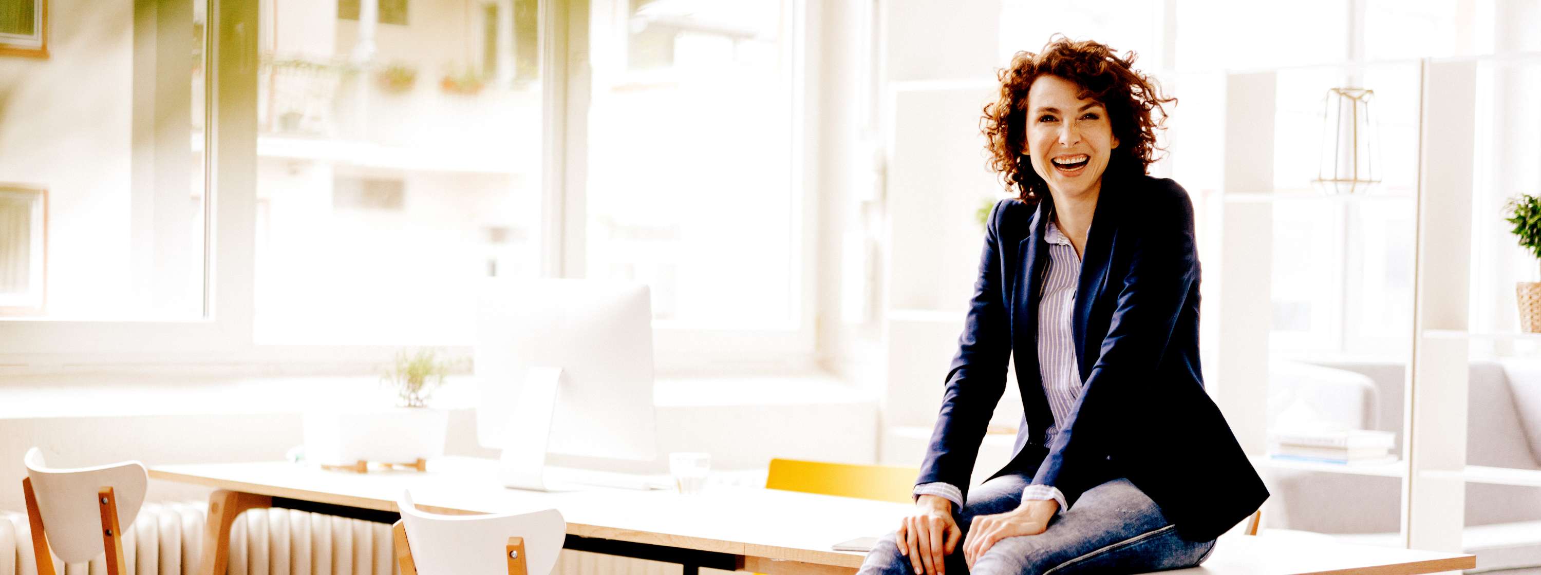 Smiling woman with light tone skin and short brown hair sits on the table in a sunny boardroom.