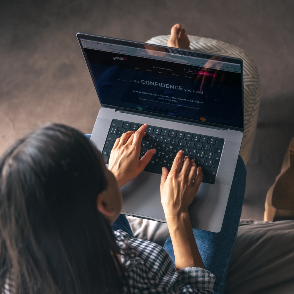 A dark-haired woman working on her laptop. The Qtrade Direct Investing website is displayed on screen. 