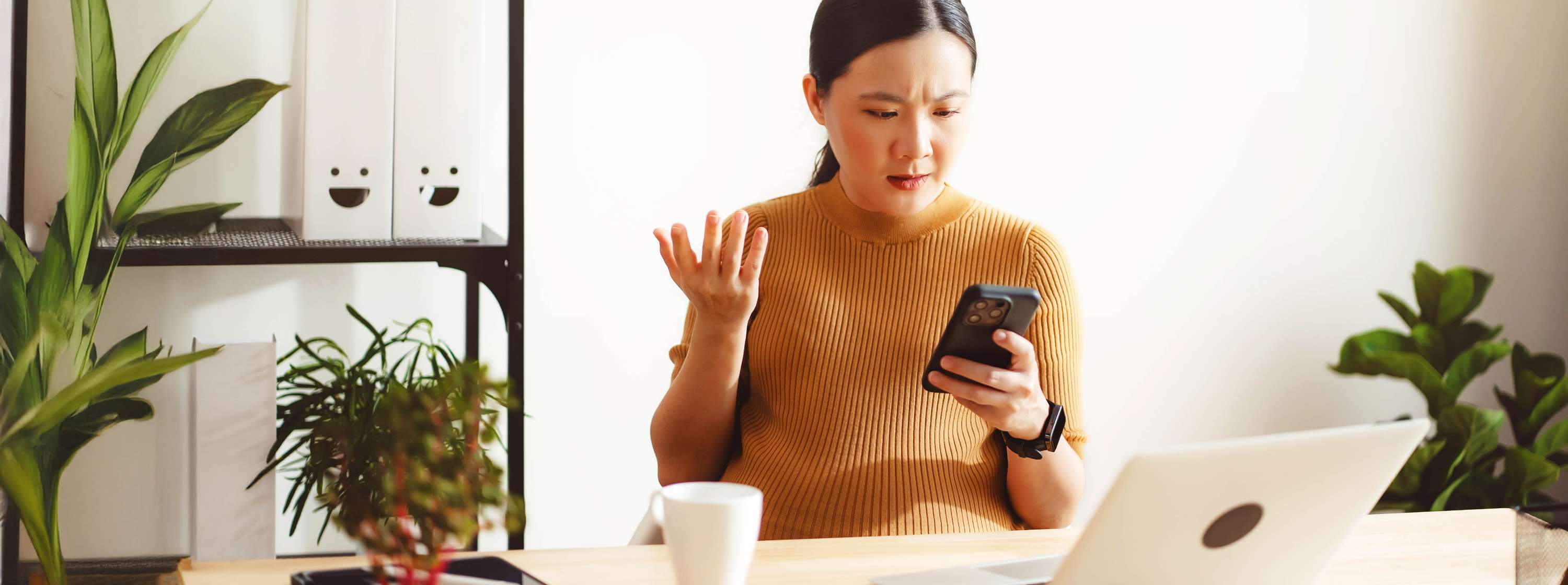 A young woman wearing a mustard-colored sweater is sitting at a desk, with a frustrated look on their face, holding a smartphone in one hand and gesturing with the other. On the desk, there is a white coffee mug and an open laptop. In the background, there are shelves with white storage boxes featuring smiley faces and a potted plant.